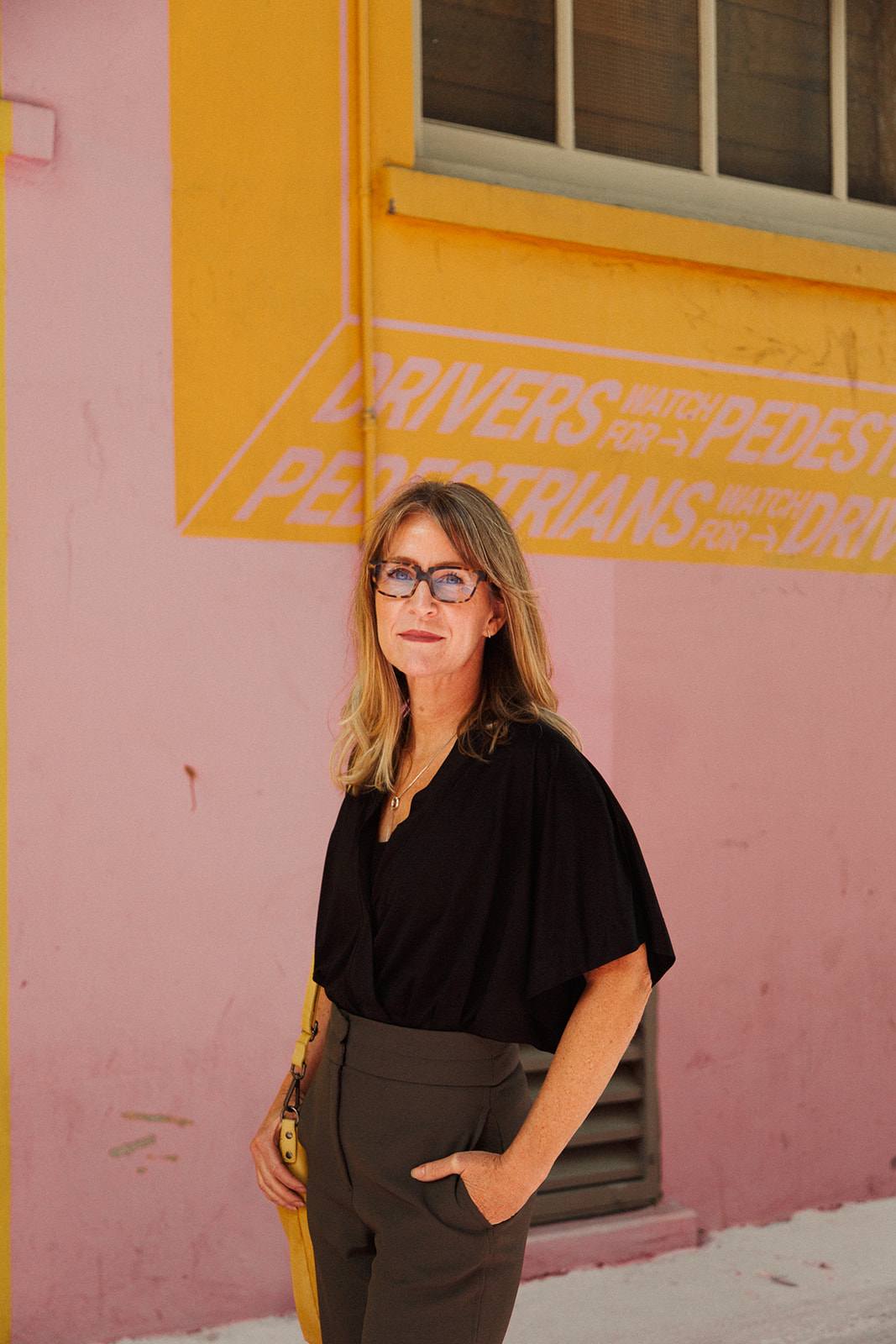 Portrait of Melissa Higgs. She stands in front of a pink and yellow painted wall (found within the Alley Oop laneway in downtown Vancouver).