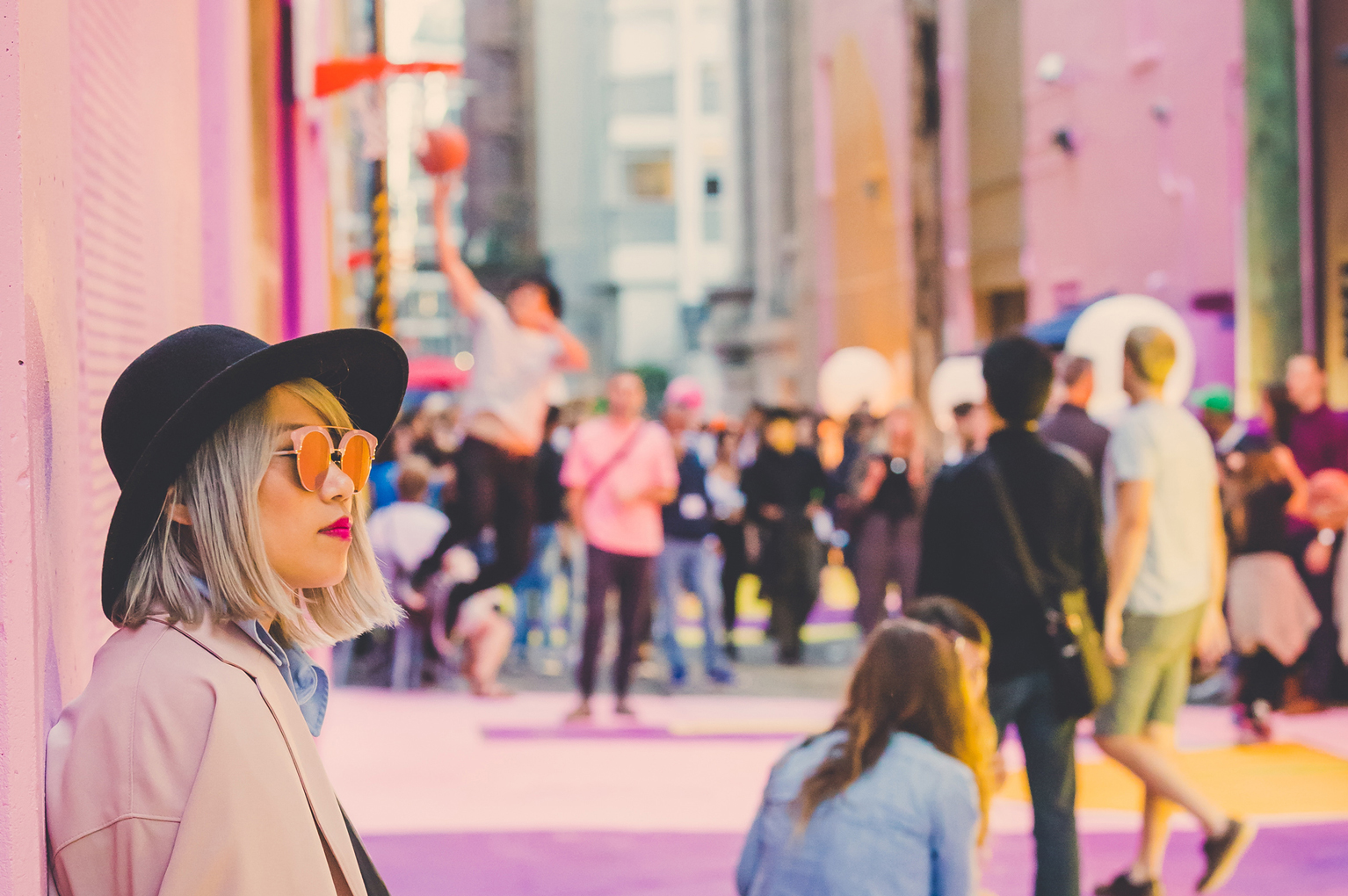 A woman stands against a pink wall in a downtown laneway. The laneway is fully painted, walls included, in pink, purple, and yellow, with lines for a basketball court. Blurred in the background, the laneway is bustling with activity, with some people playing basketball with a hoop attached to the wall.