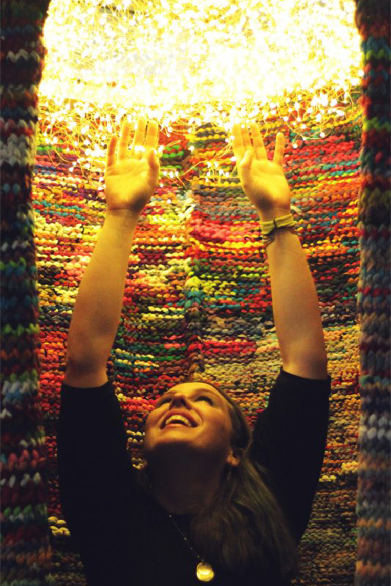 Woman sitting in the finished piece with her hands raised to touch the fairy lights.