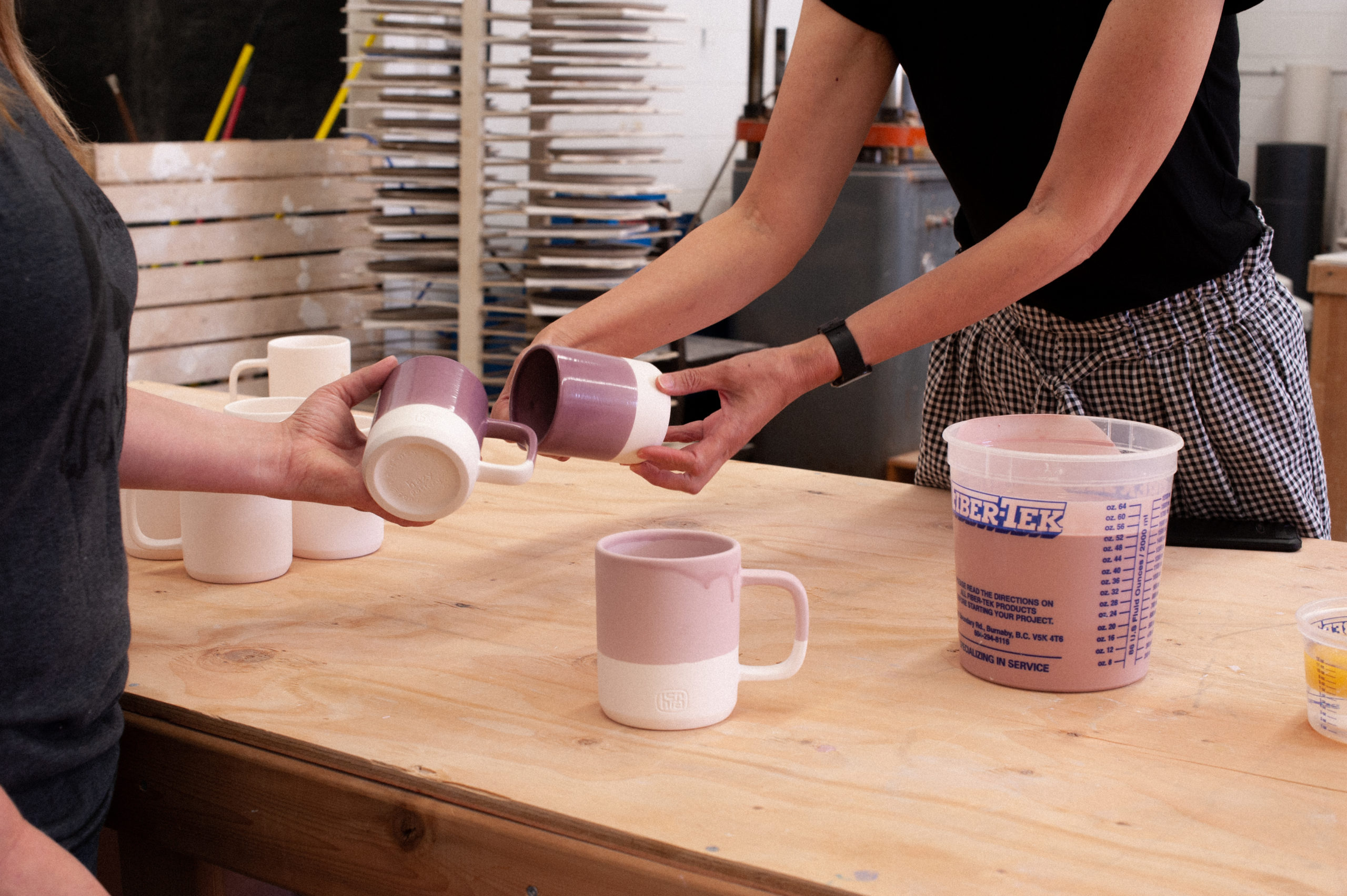 Mindy Doke and colleague holding HCMA mugs glazed in eggplant across a wooden workbench with a tub of glaze and unfired mugs scattered around.