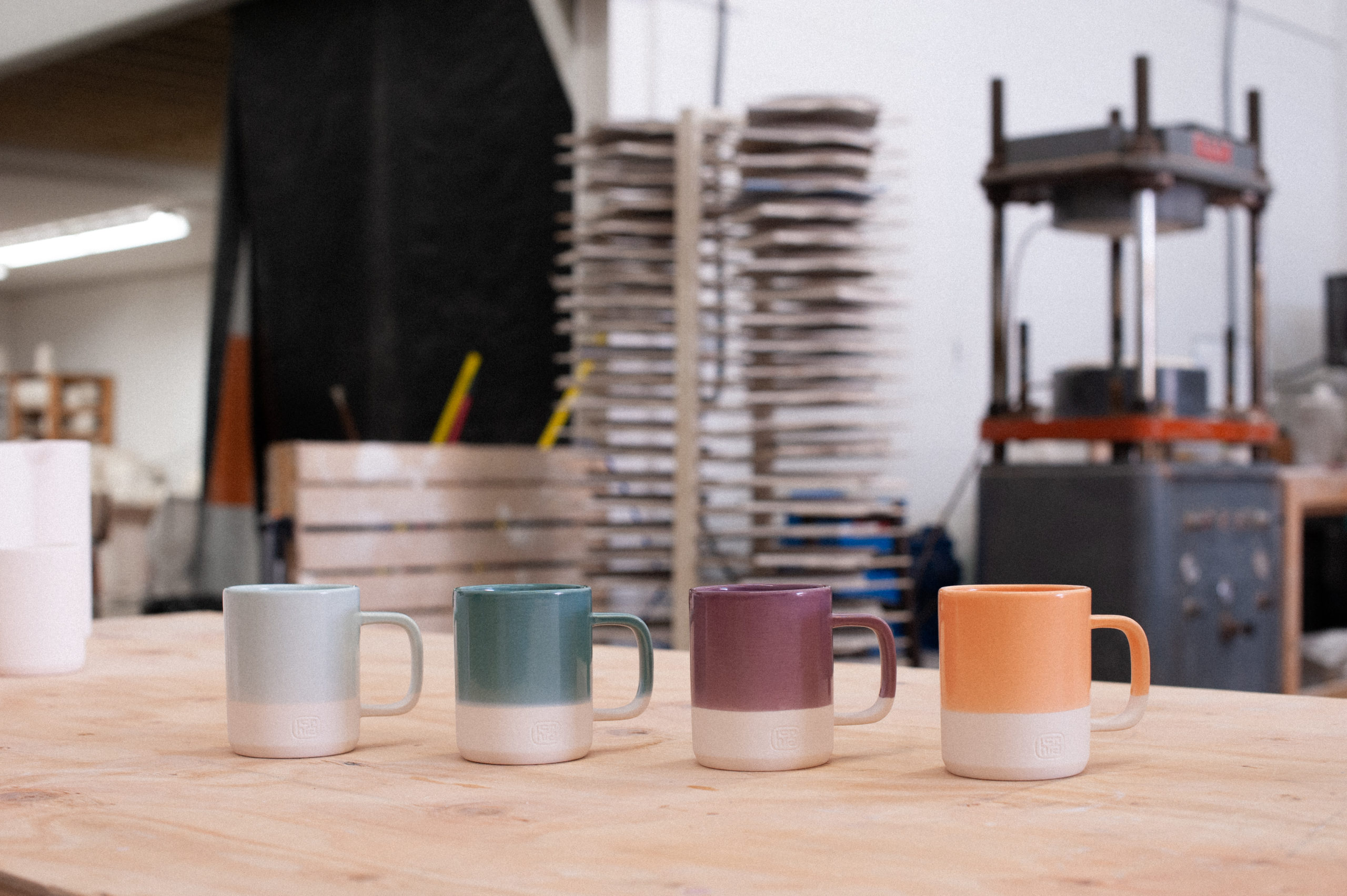 Three finished hcma custom mugs, freshly glazed in 4 colour-ways, light blue, team, eggplant and orange. They sit on a wood-topped work bench in the hazy pottery studio.
