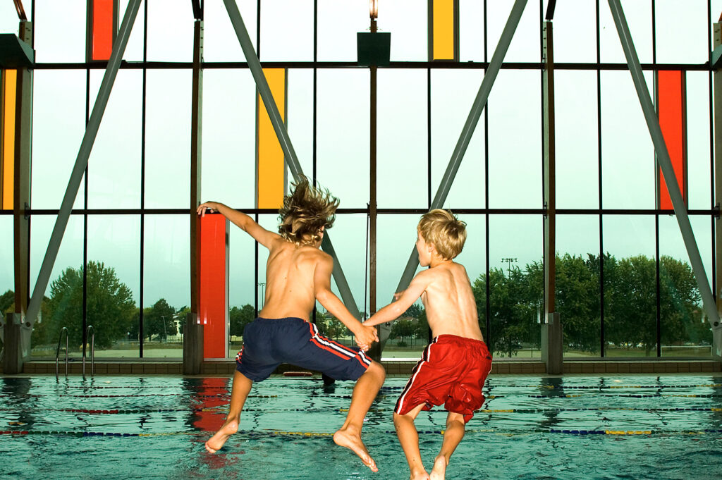 Two little boys jumping into the pool with a large window in front of them.