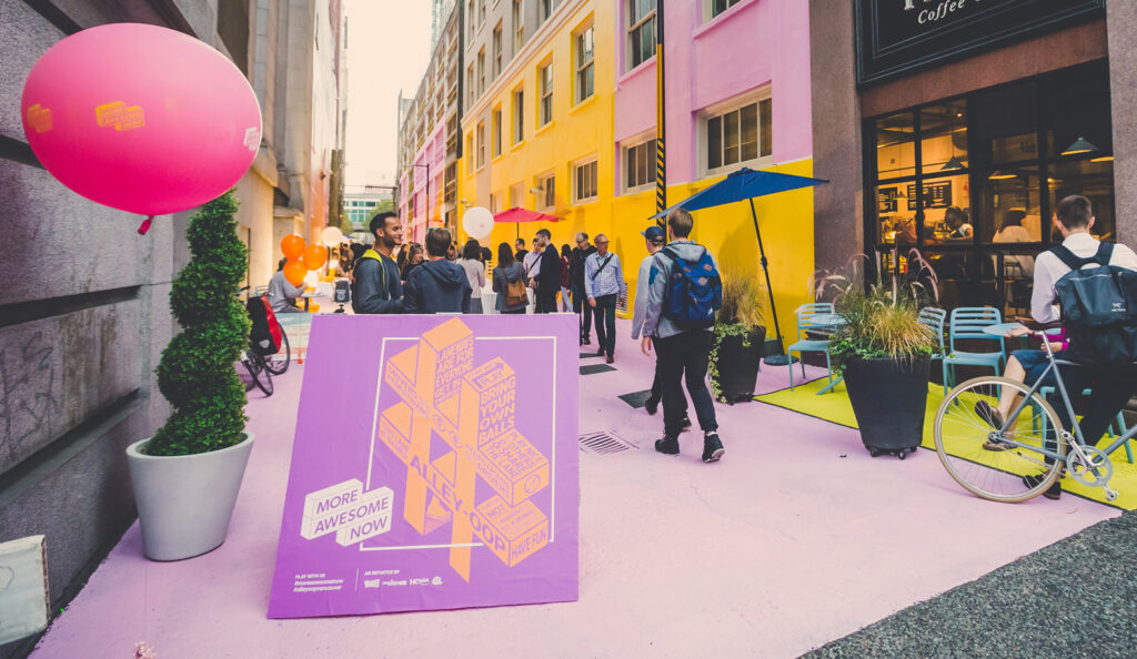 People gathered in Alley-Oop, a viibrant laneway activation. There is a sign in front with the "More Awesome Now" logo, and a 3D hashtag graphic.