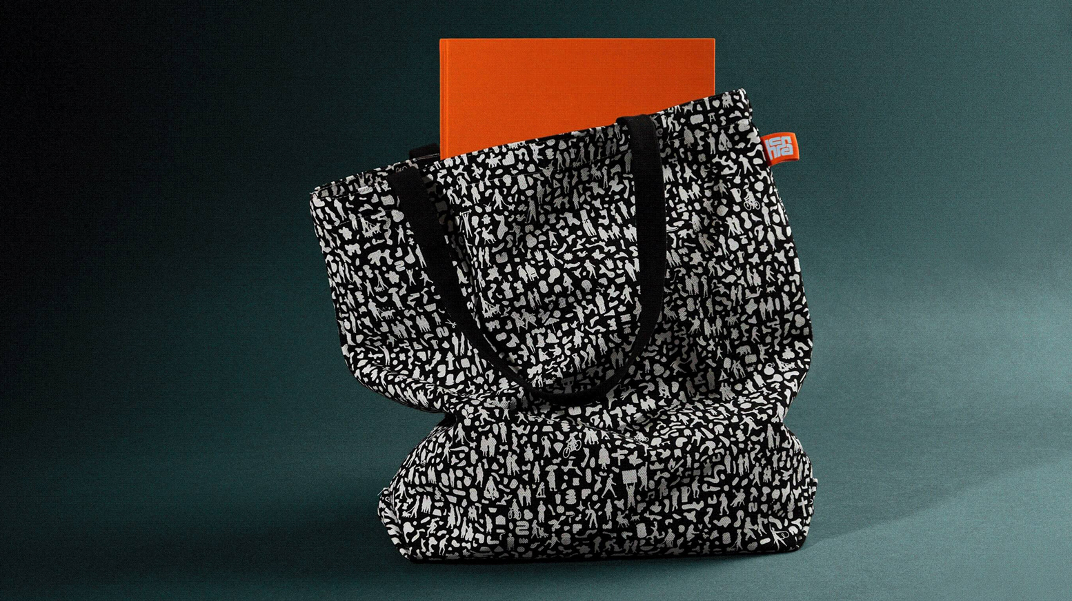 HCMA branded tote bag with a red book sticking out of it. The bag is black with white silhouettes of people and objects shrunken down and tightly squeezed together as a pattern.