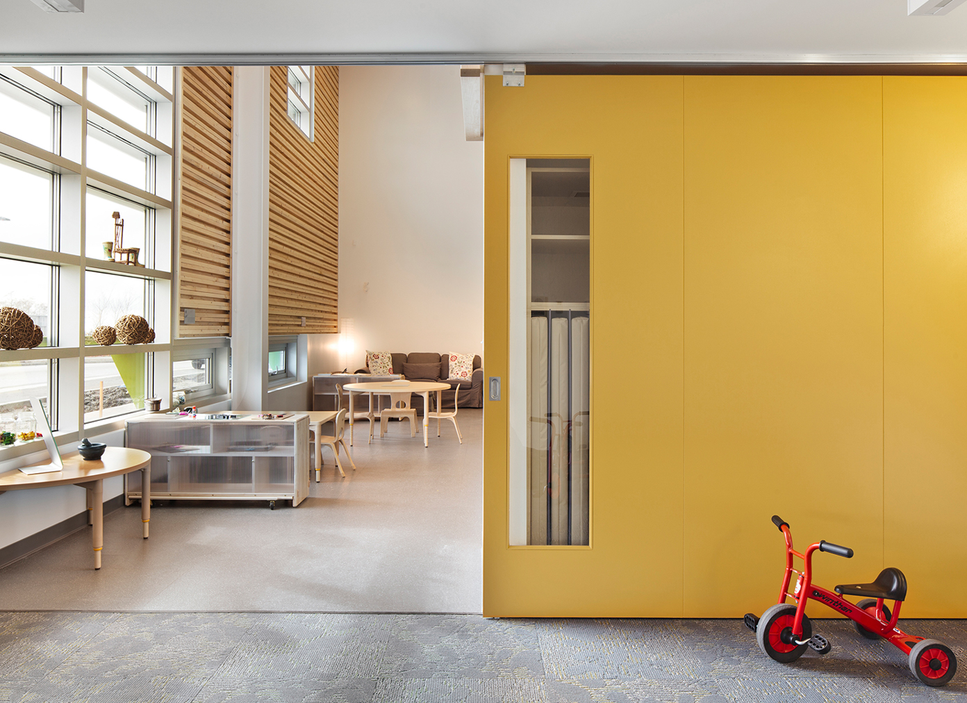 A red tricycle sitting in front of a yellow wall that leads to a classroom with wooden tables and chairs