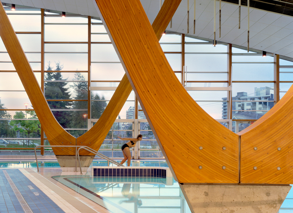 The West Vancouver Aquatic centre displaying someone jumping in between large wooden pillars in the shape of an organic 'V'.