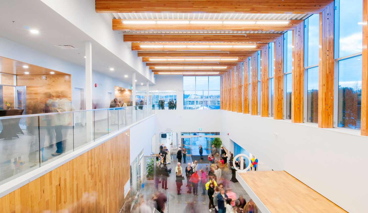 Interior view looking down from the second level of Timms Community Centre with people. The combination of natural, bright white interiors, and large windows make the space feel open and welcoming.