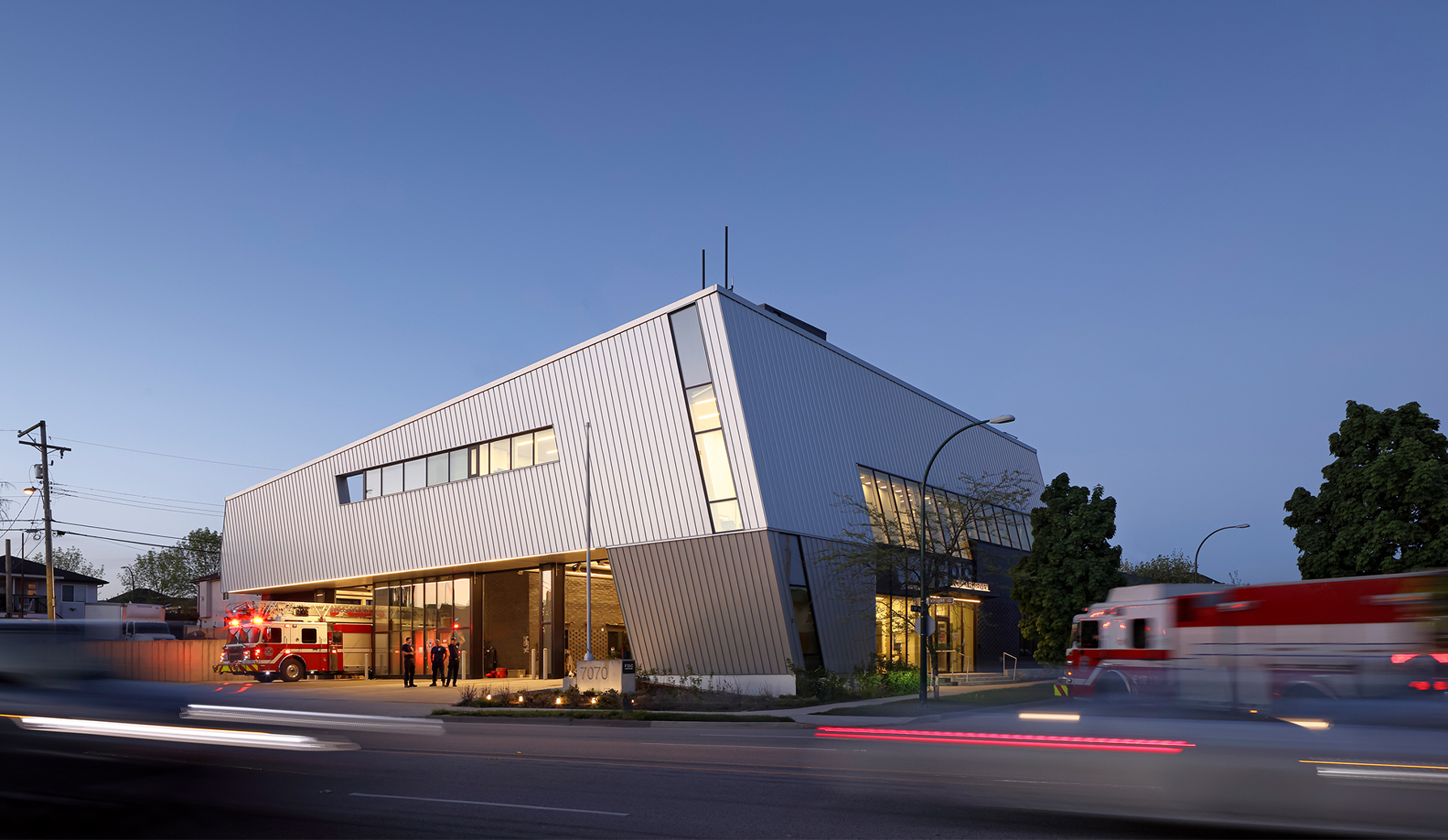 The Vancouver Fire Hall 17 exterior as viewed from across the street, at dusk. The sky is a fading periwinkle while blurred cars whizz by creating streaks of red. A fire truck is moving out of one of the bays with its lights on, while another on the road moves towards the facility. A group of firefighters are standing on the concrete apron outside.