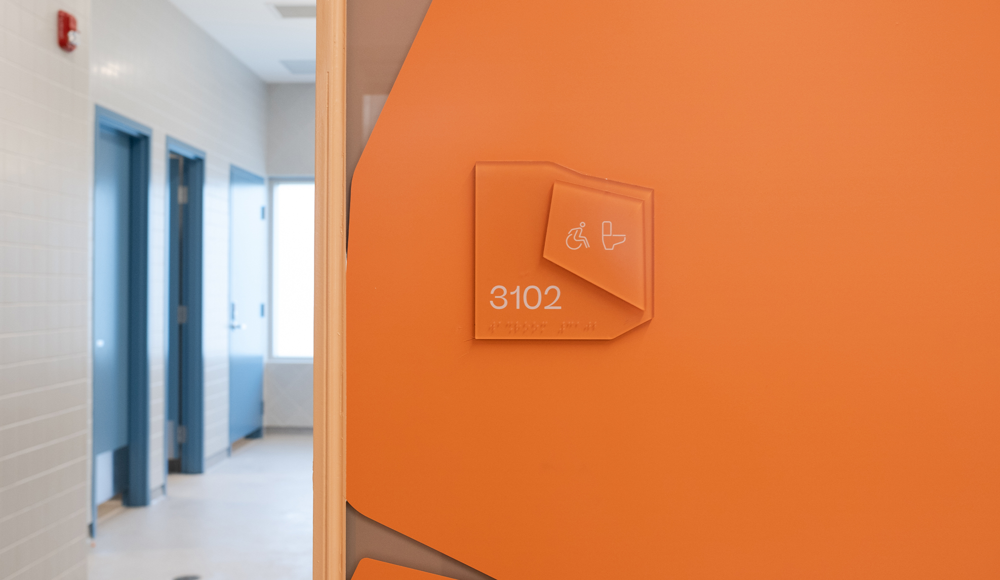 An orange wall sign on an orange wall in a rock-like shape that says "3102" and displays a wheelchair and toilet icon.