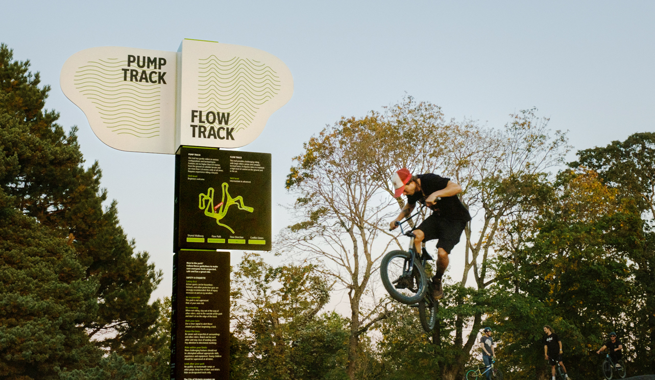 A tall sign that has an organic-shaped sign on it that says "FLOW TRACK" with a map. A biker is doing a jump behind the sign.