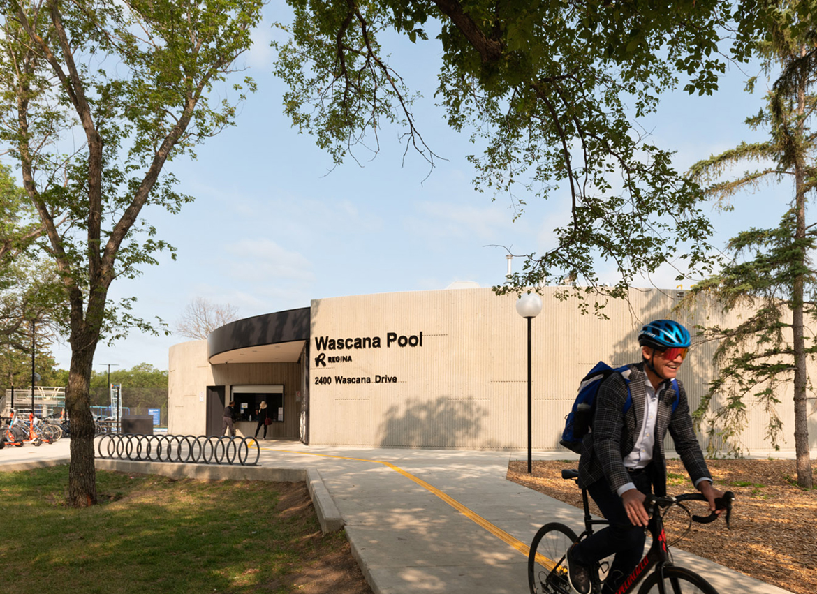 A man bikes past the exterior of the Wascana Pool. Trees are shading the area. In the background, two people stand at the entry window.