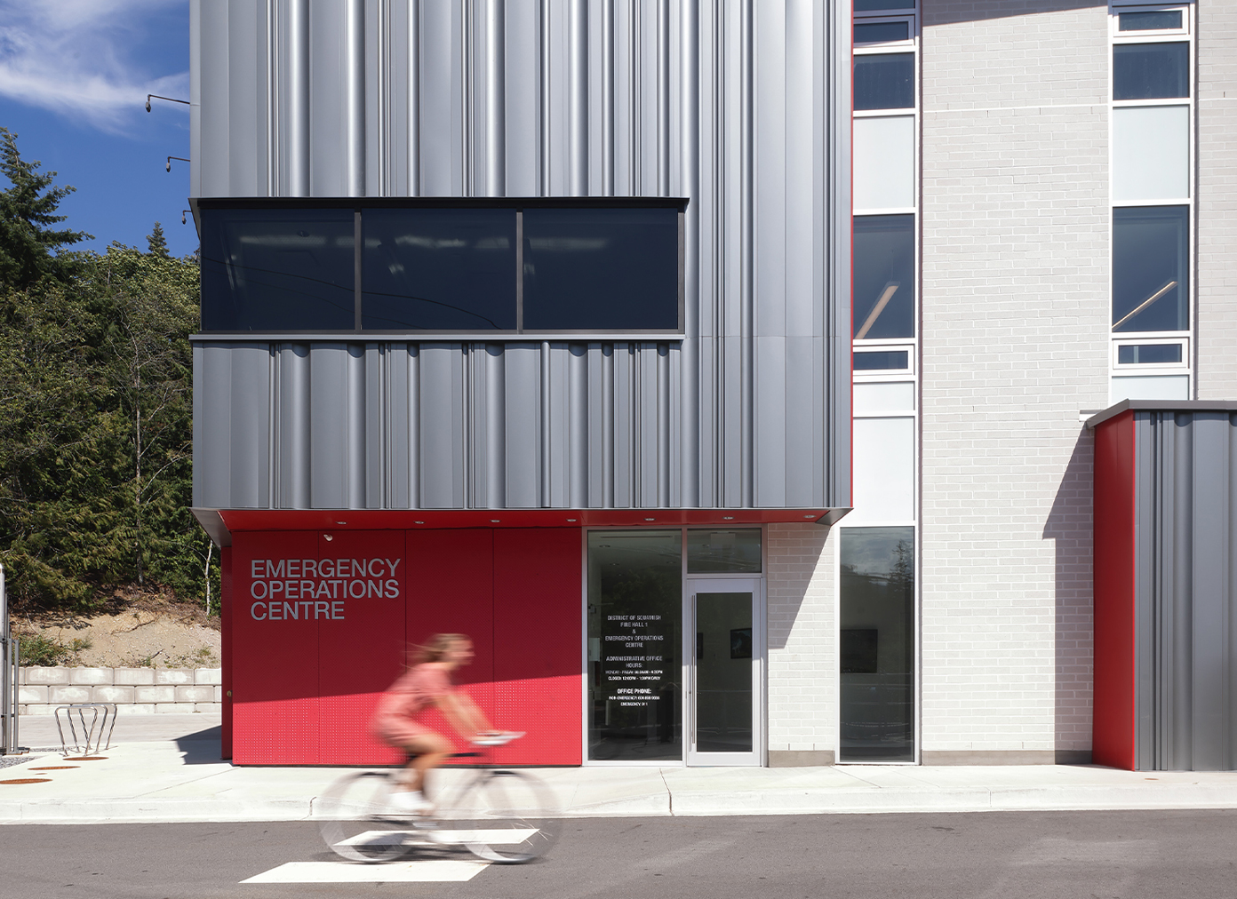 Someone biking past the Emergency Operations Centre in Squamish Fire Hall 1. The building embraces its pops of red paint and seemingly metal panelling.