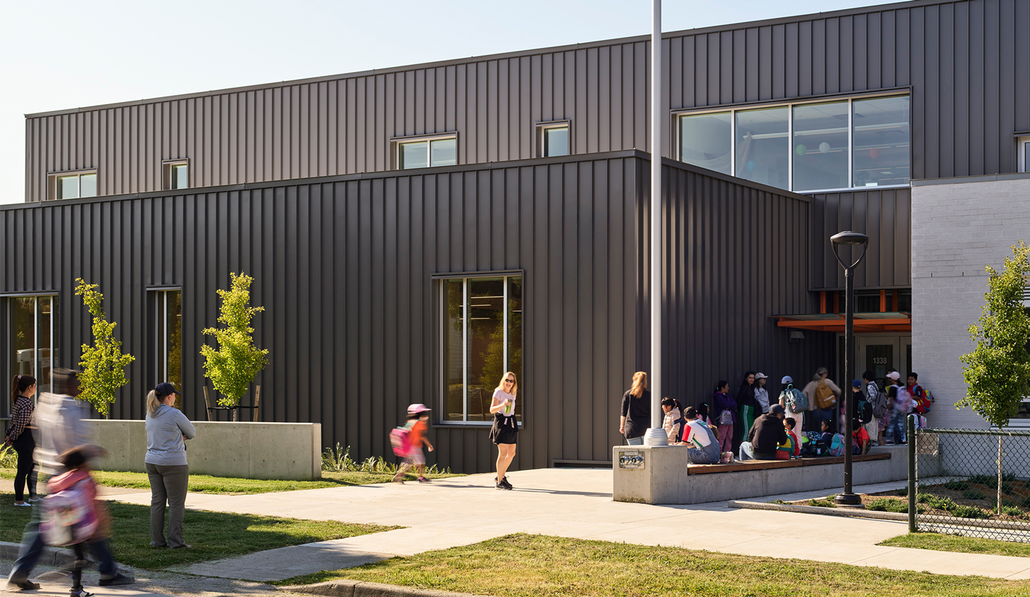 Photo of the David Lloyd George exterior on a sunny day. Students are pictured filing inside which adds colour and movement. The school has a dark grey metal cladding paired with clean white brick.