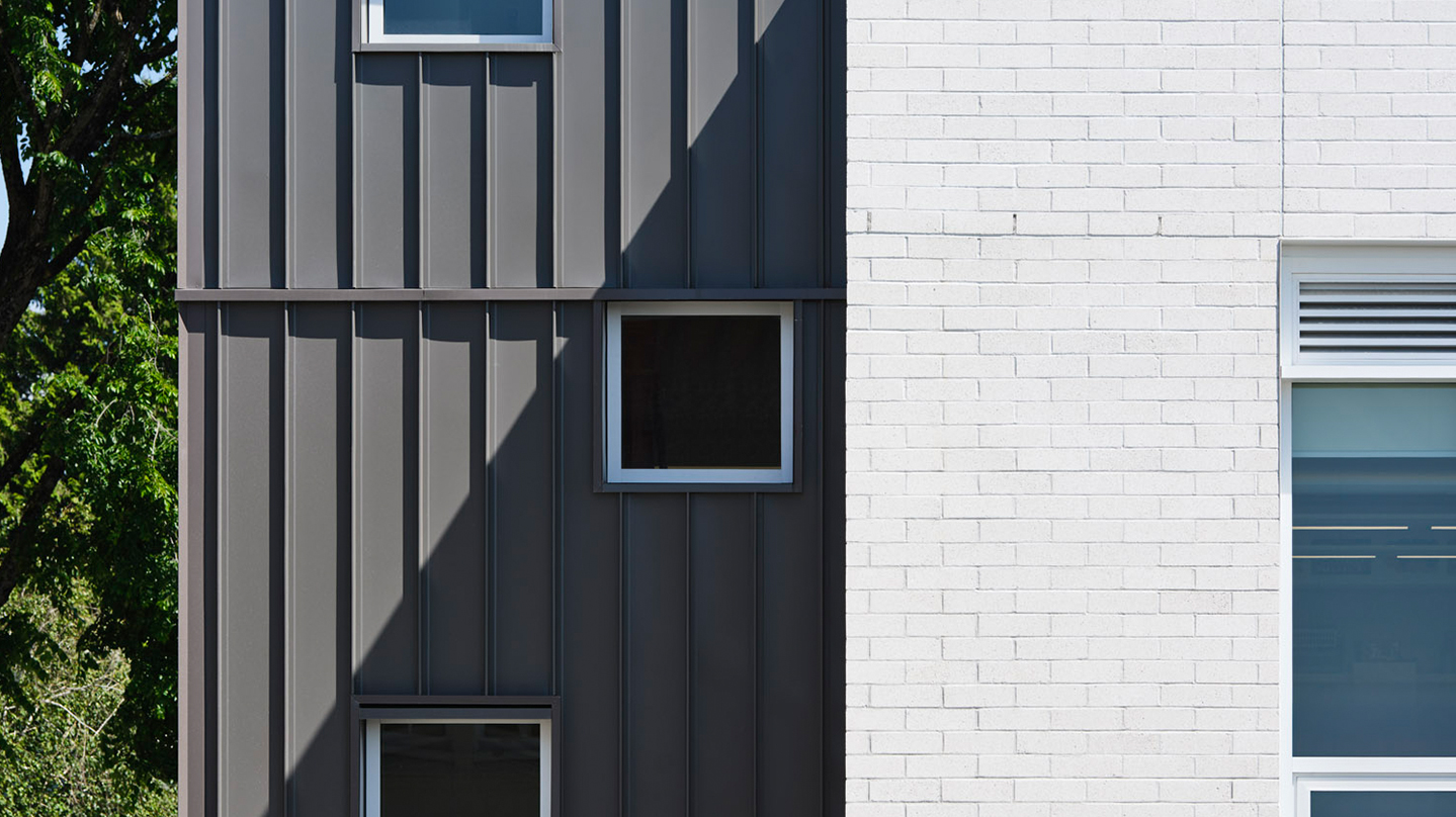 Close-up of the school's facade showing a contrast between dark vertical metal cladding with small square windows on the left, and white-painted brick with a larger rectangular window on the right. A diagonal shadow cuts across the cladding, adding depth and visual interest. Trees are partially visible in the background.