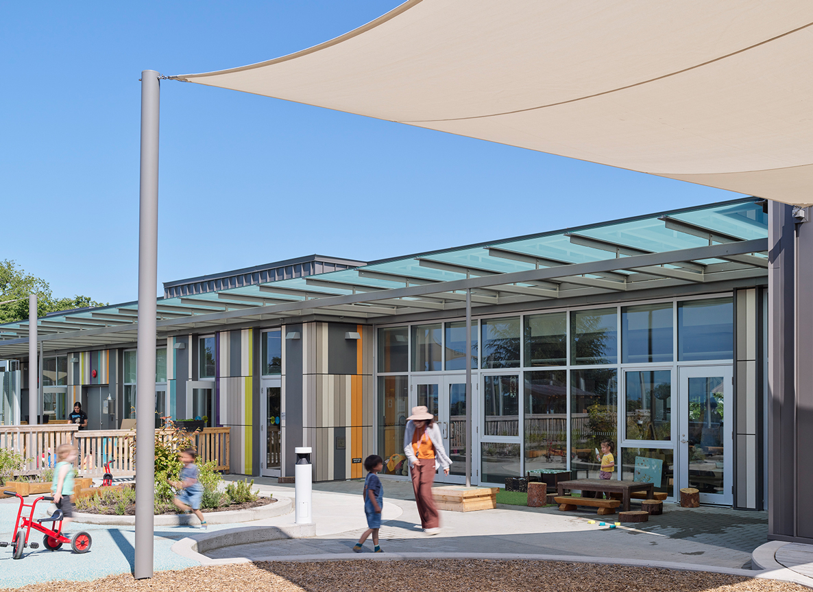 Rooftop childcare facility at David Lloyd George Elementary School, featuring a modern design with floor-to-ceiling windows and colorful vertical paneling. A glass canopy extends along the facade, providing shelter over the entrance and outdoor play area. Children play with toys and tricycles while a caregiver supervises. A large tensile fabric shade structure offers sun protection, and landscaping elements soften the rooftop environment under a clear blue sky.
