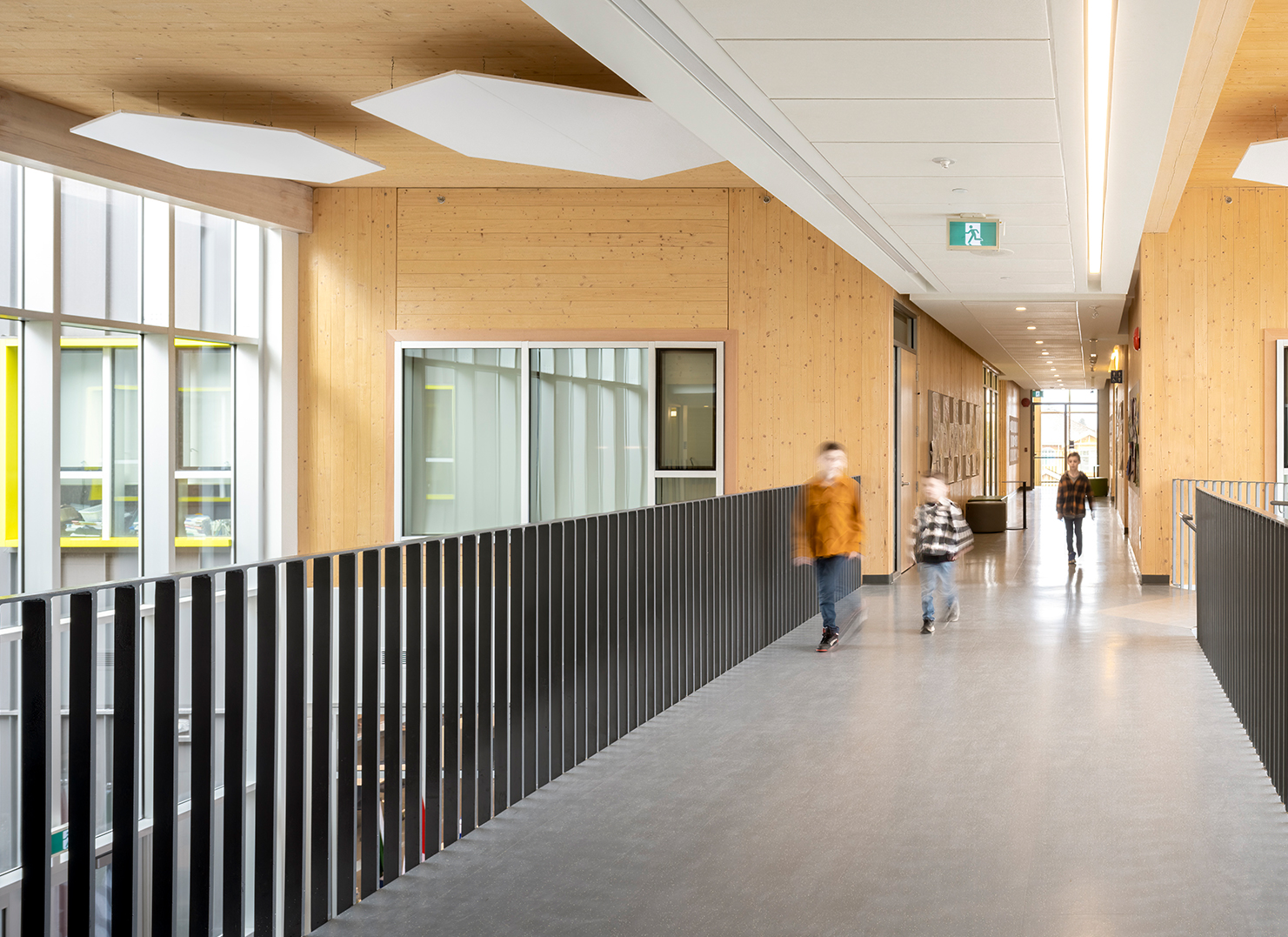 A long, well-lit corridor in the school, with wooden walls and ceiling in the background and a metal railing on either side of the walkway. White hexagonal panels are attached to the ceiling. Blurred figures are passing through.