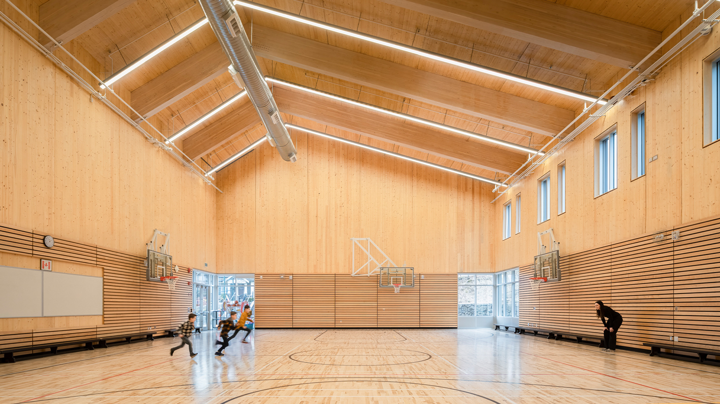 A spacious gymnasium with high wooden ceilings and natural light streaming through the windows, with students playing a game. The gymnasium is entirely wooden, from top to bottom. Children run from one side of the gym towards an adult on the other side.