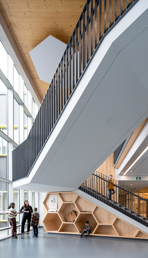 A modern school hallway featuring a staircase with integrated hexagonal wooden cubbies and seating areas where students interact.