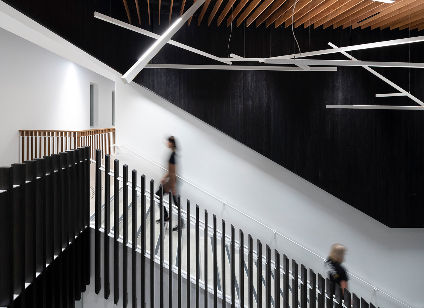 Blurred figures walk up a central staircase. Above, a feature ceiling with wood slats and multiple long, rectangular pendant lights.