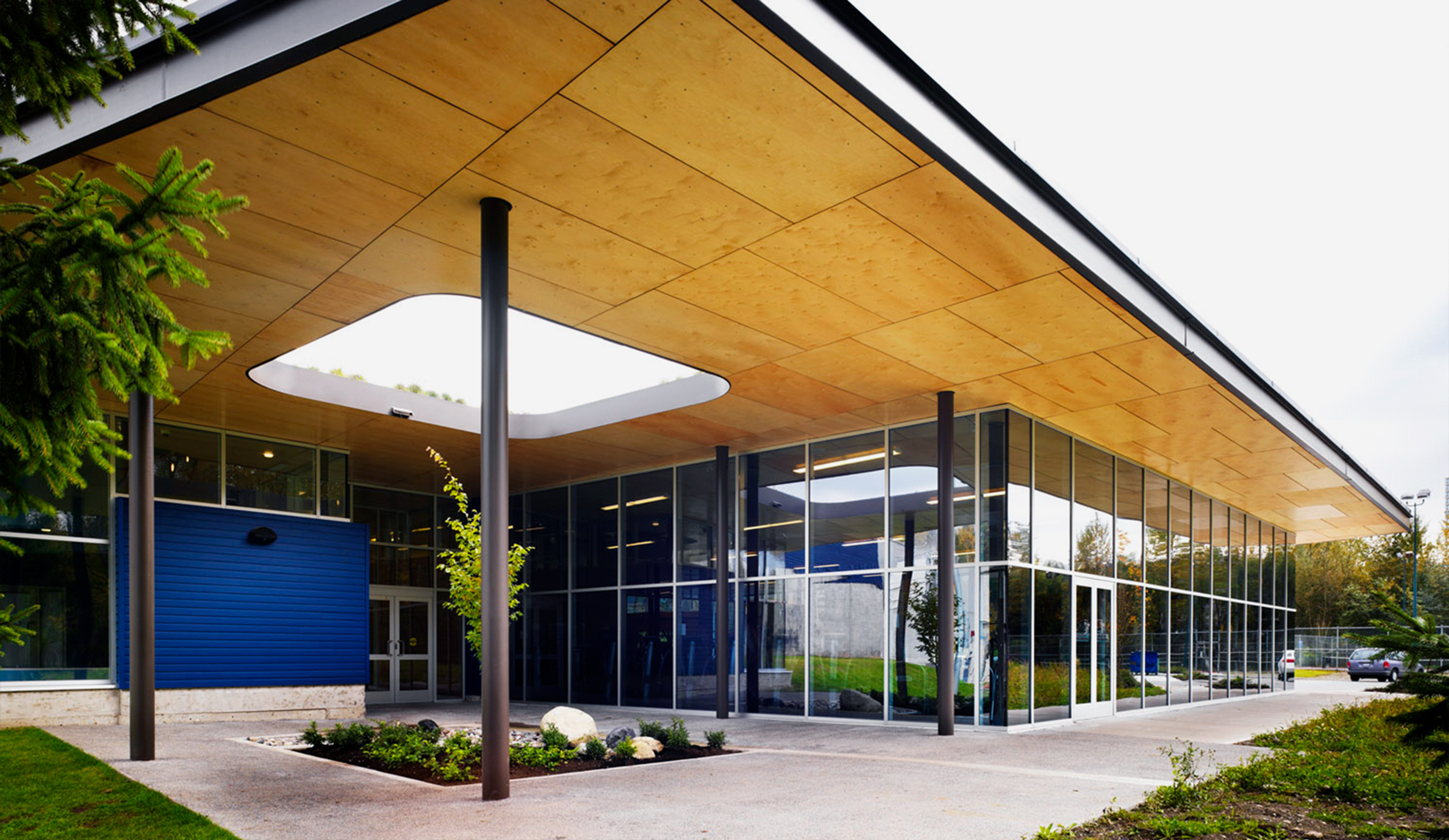The front entrance of Port Moody Recreation Centre. It showcases a wooden overhang with a rounded square skylight, leading to a garden below.