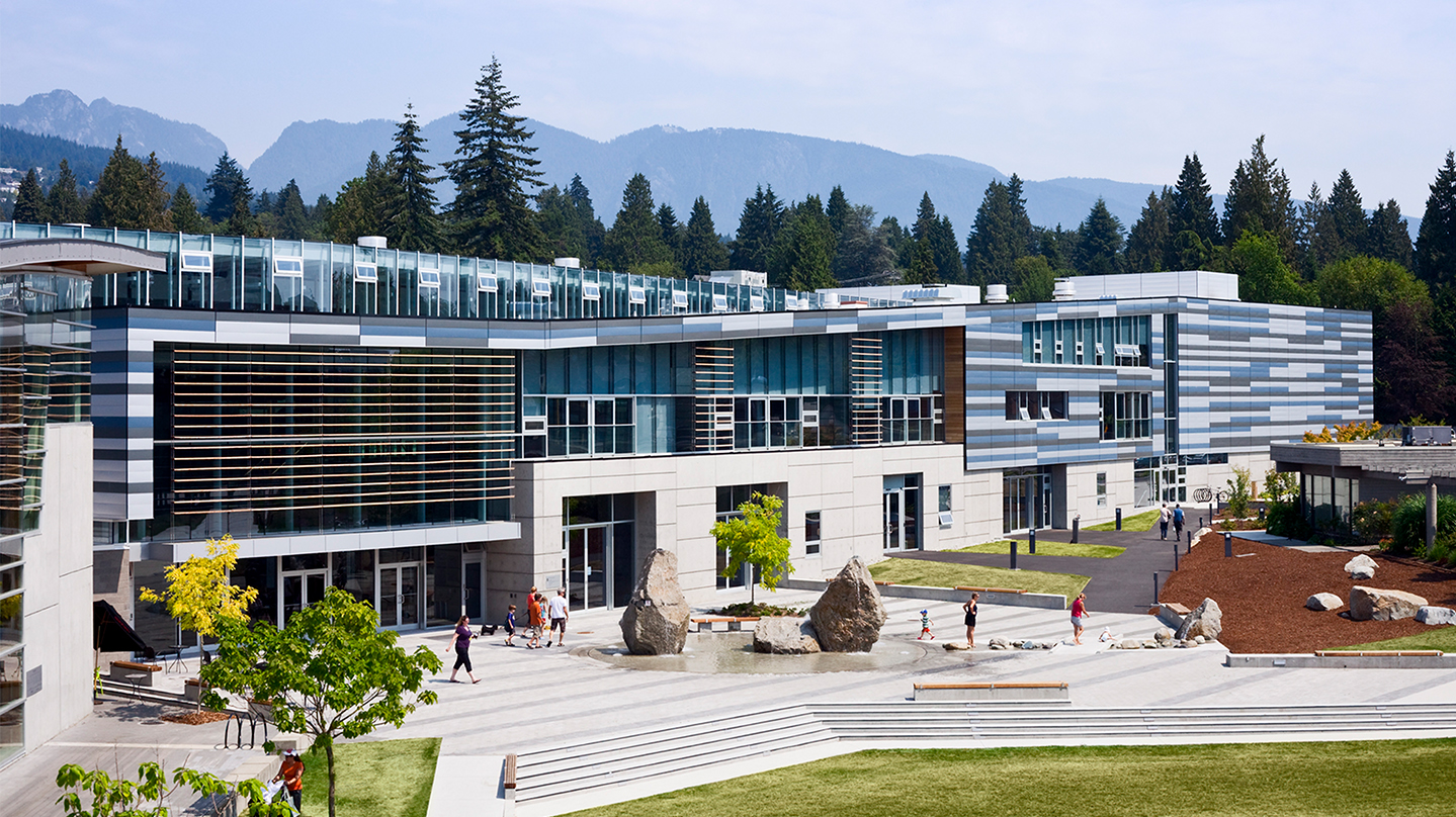 Exterior of the West Vancouver Aquatic and Community Centre, with the mountainous, forested context in the background, and the great lawn stretching into the foreground. Various patrons mill about outside.