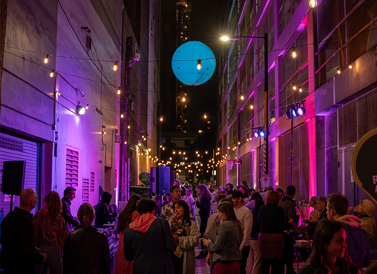 Alley Oop at nighttime during a fundraising event. People fill the alley while purple and pink lights flood the walls and twinkly lights hang above them.