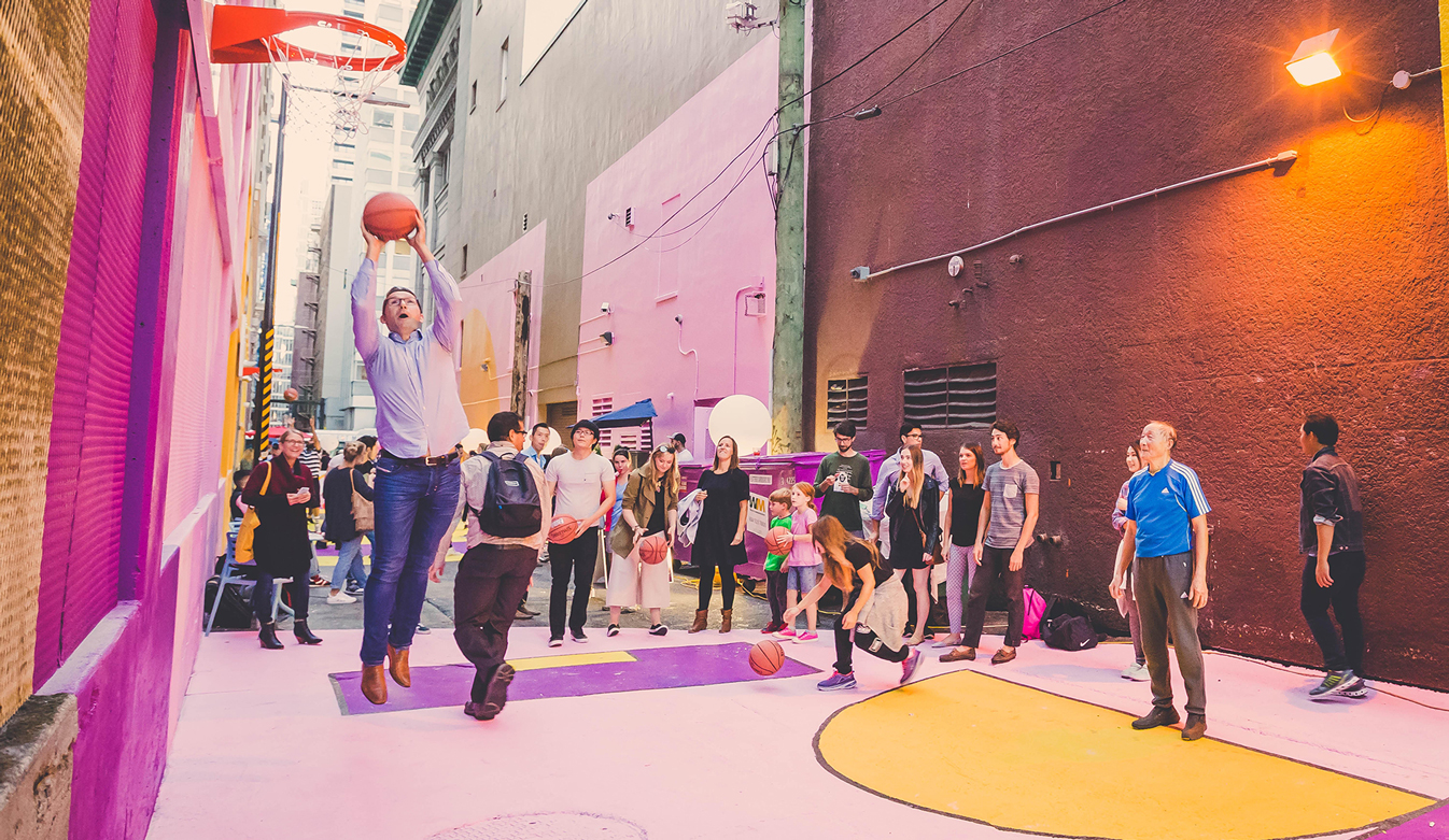A man shoots a basketball at a hoop installed on the wall within Alley Oop while a large group of people gather around him. The alleyway ground and walls are painted pink and yellow.