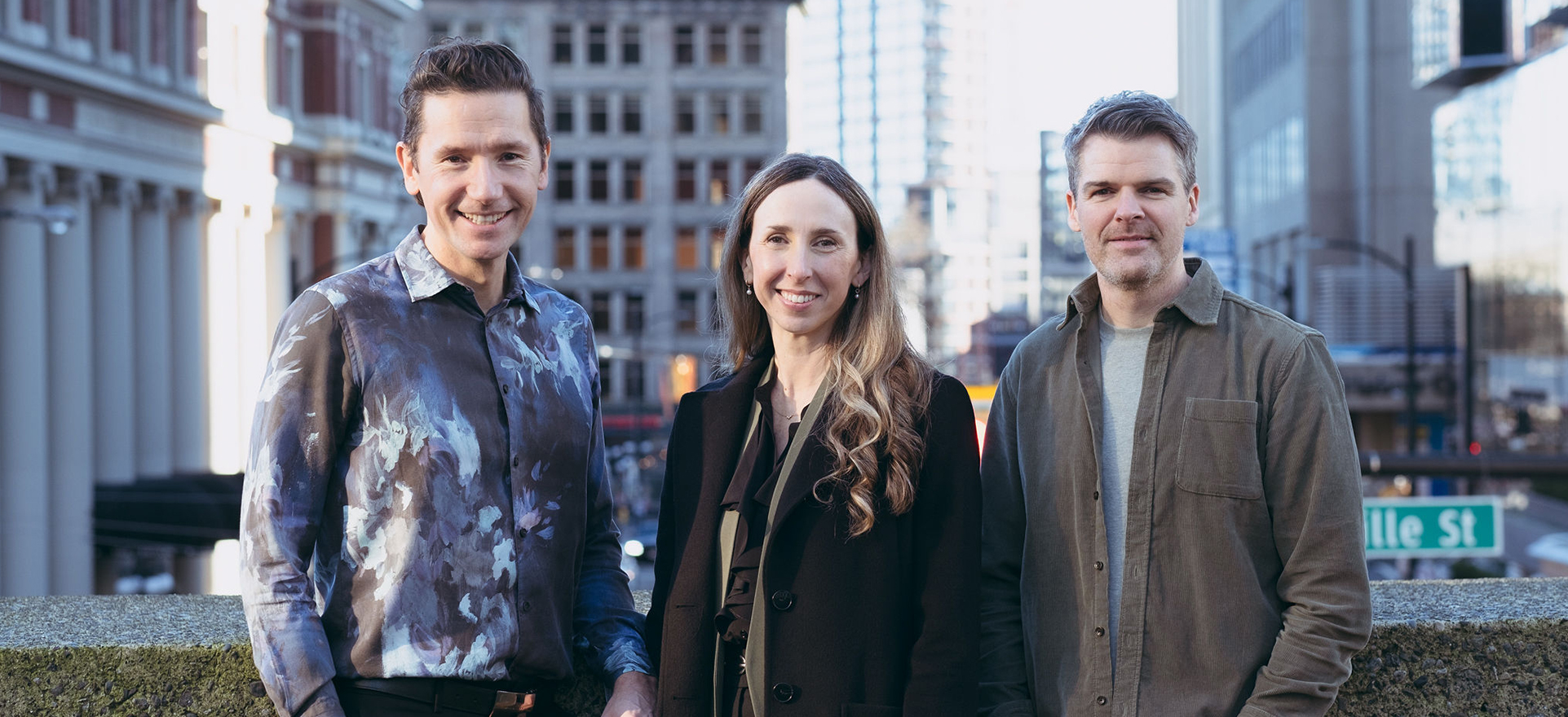 From left to right, Aiden, Ali, and Darin stand together smiling at the camera. They lean against a short concrete wall, and in the background behind them is a slightly blurred view to Granville Street in Vancouver.