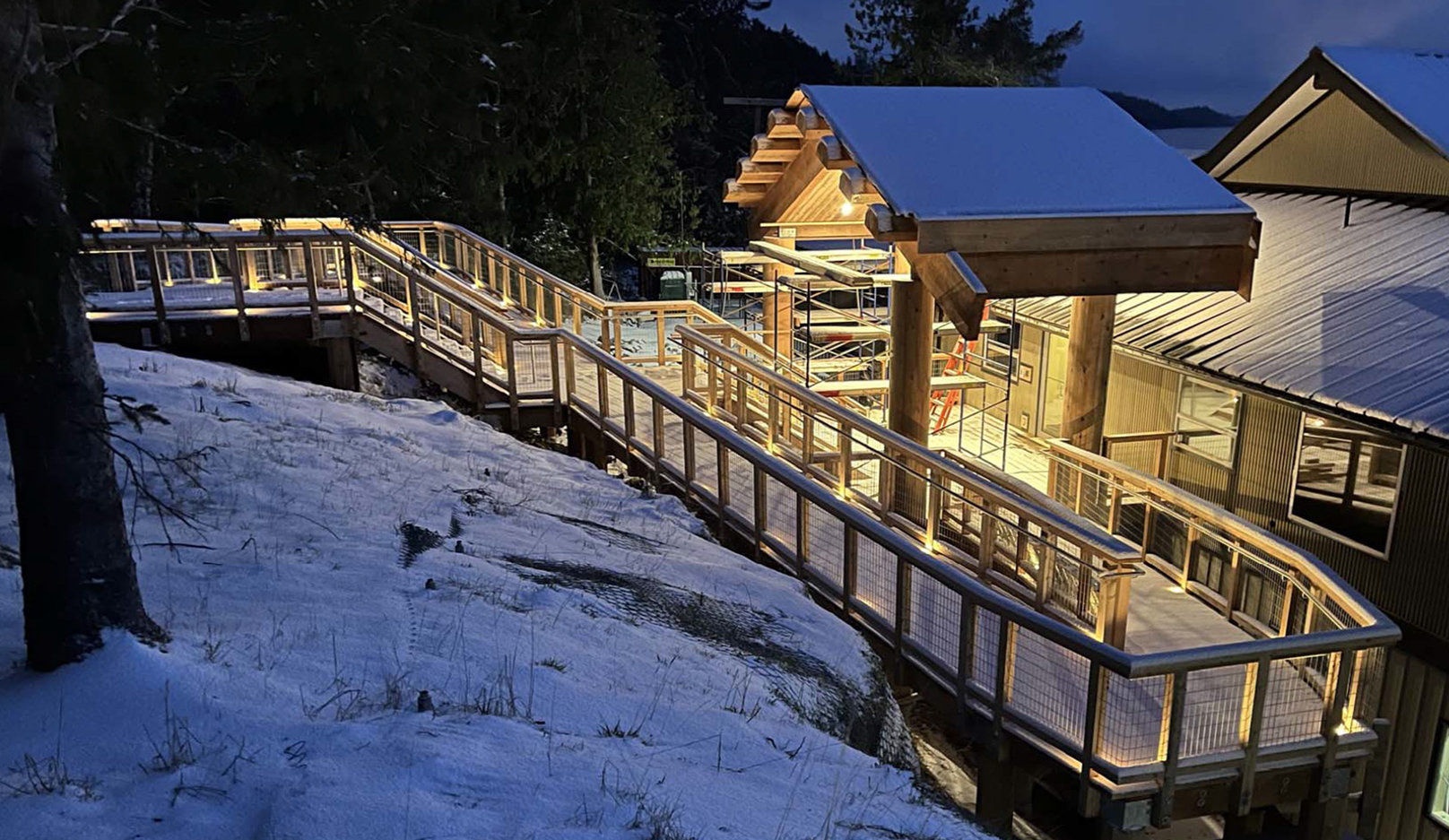 Photograph of new entranceway during construction. The image show the new accessible ramp and stairs in the foreground, connecting to the new covered exterior entranceway canopy. The canopy and ramp is of cedar construction with large locally sourced timbers. Roof timbers were carved locally to resemble traditional Haida Gwaii longhouse forms.