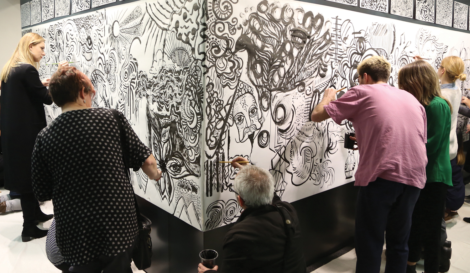 Photo from Ola Volo's Sumi Ink workshop, with people painting onto large sheets of paper pinned on the Vancouver office's magnetic wall. It features an eclectic mix of abstract patterns, faces, eyes, and organic shapes. Each person is focused on painting their section using brushes and black ink.