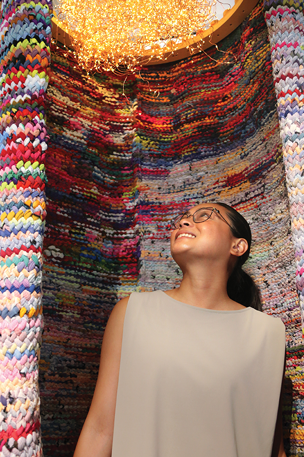 An hcma-er stands within Katherine Soucie's piece, Cast ON, Cast OFF, a tall, tubular, woven piece knit from hosiery in various bright colours. She looks up at a light fixture at the top of the piece that glows warmly.