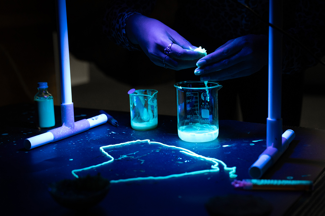 Hands play with glowing slime within a glowing beaker, captured from Marni Bowman's residency, Play in Process.