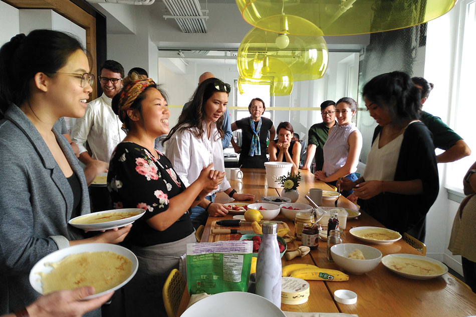 A group of people stand around a table in the kitchen at hcma's Vancouver office, making crepes as part of one of Annabelle Choi's residency workshops.