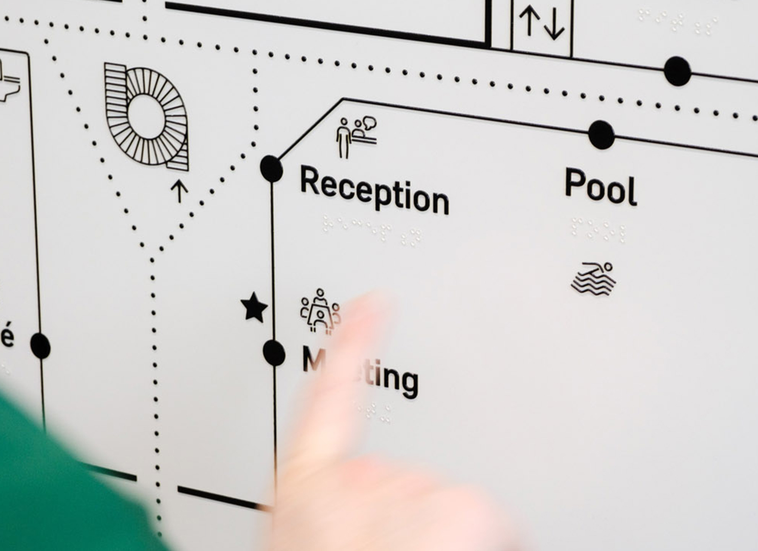 A blurred hand touches a tactile floorplan of the facility's first level.