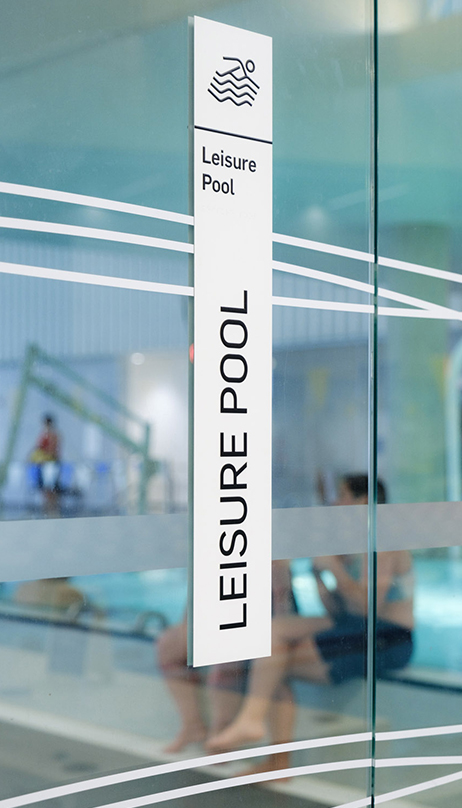 Signage for the leisure pool paired with visibility graphics on a glass wall. Through the glass, blurred figures are sitting on a transfer ledge into the leisure pool.